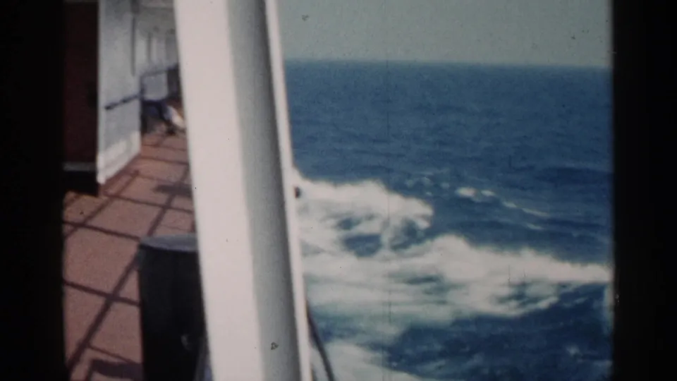 The frame shows a steady 1960 8mm deck perspective with a white rail, reddish deck, and foamy wake trailing across blue water.