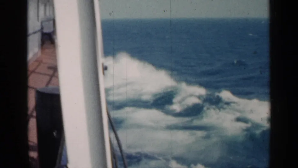 A shipboard 8mm frame shows rolling white wake, a vertical white rail, blue open water, and soft archival color fading.