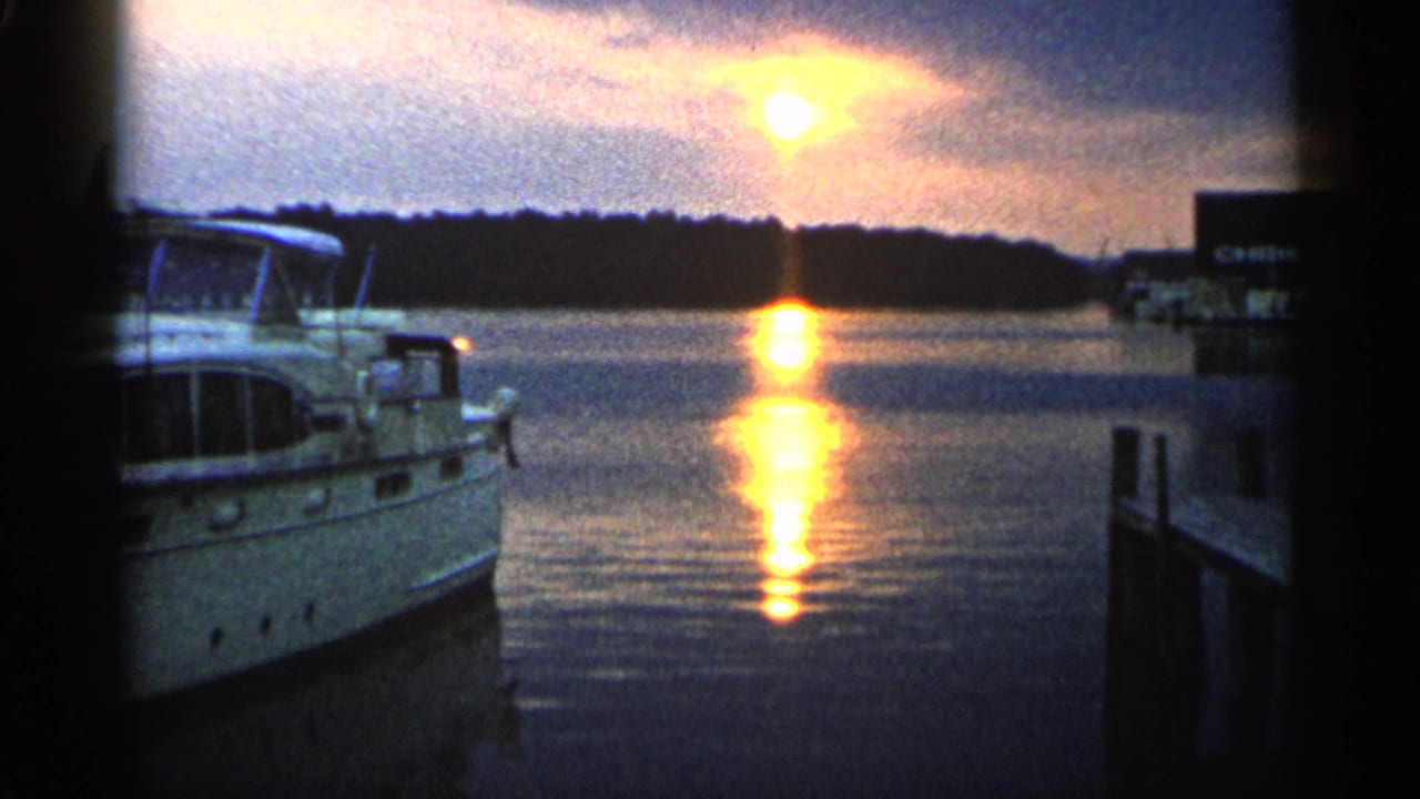 1960 Michigan harbor view with a white motorboat at left, bright sun above a dark tree line, and a vertical reflection on the water.