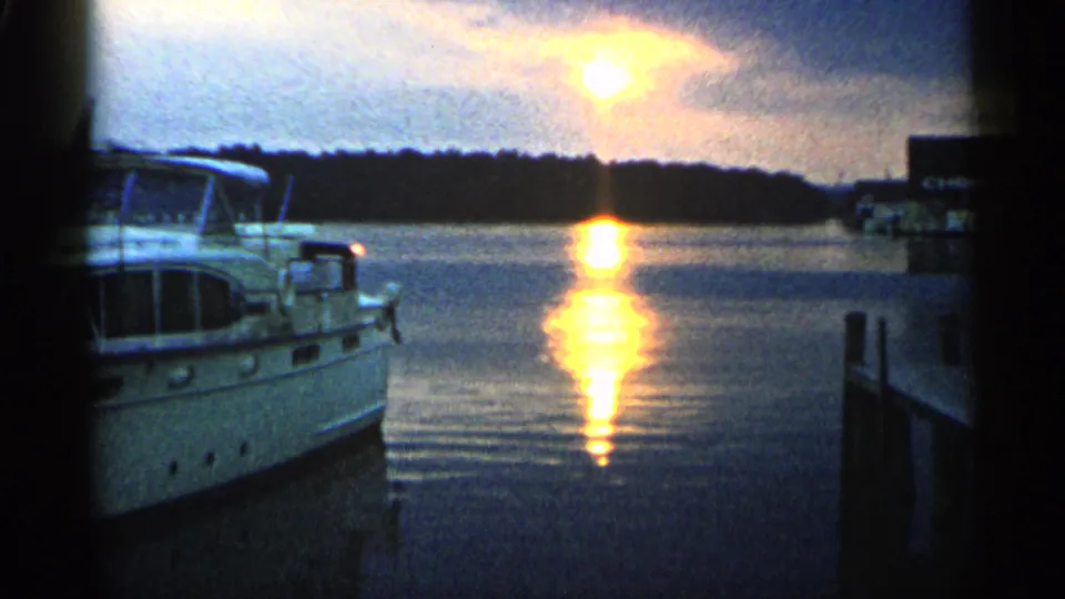 1960 Michigan home-movie frame showing a motorboat, right-side dock structure, distant tree line, and warm sun reflection on still water.