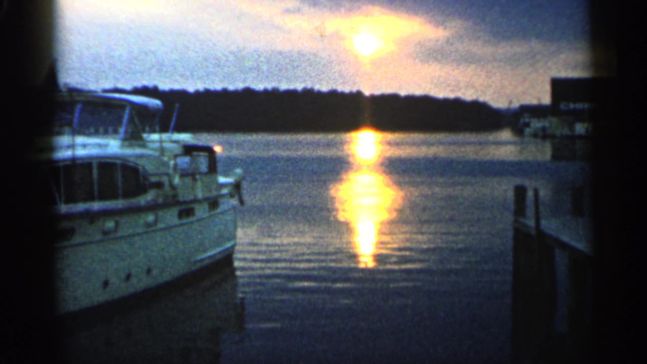 Docked white motorboat beside harbor water at sunset with treeline in the distance.