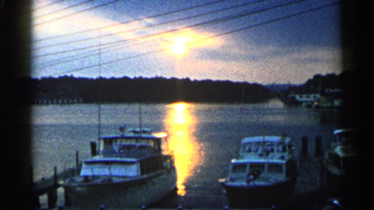 1960 Michigan harbor view with two moored boats, overhead lines, and a bright sunset path on the water.