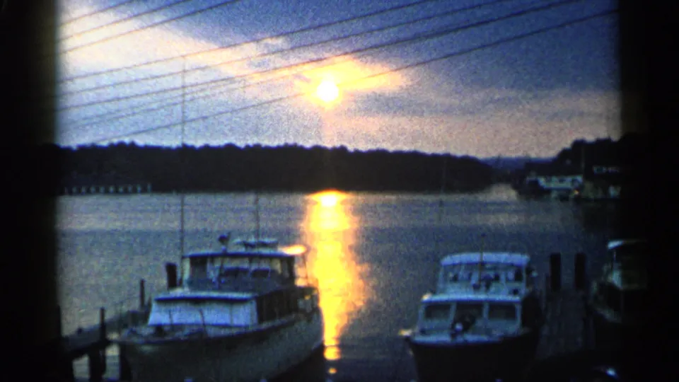1960 Michigan 8mm frame of docked cabin boats under utility lines with golden sunset reflection on the harbor.