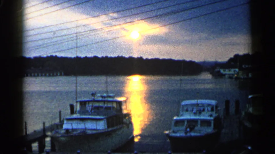 1960 Michigan home-movie frame showing moored boats, dark shoreline, and a bright sunset reflection across the water.