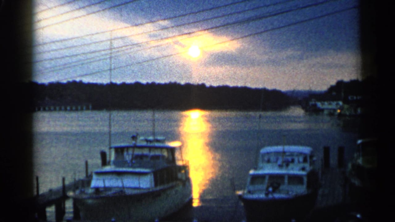 1960 Michigan 8mm frame showing docked motorboats beside a pier as the sun reflects across marina water.