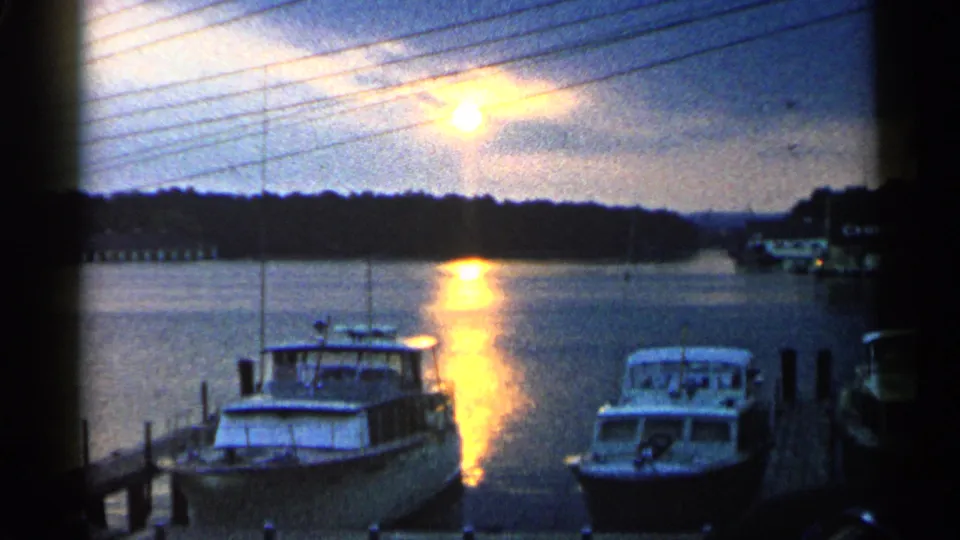 Michigan 8mm home-movie frame from 1960 showing a marina at dusk with boats, pier posts, and reflective water.