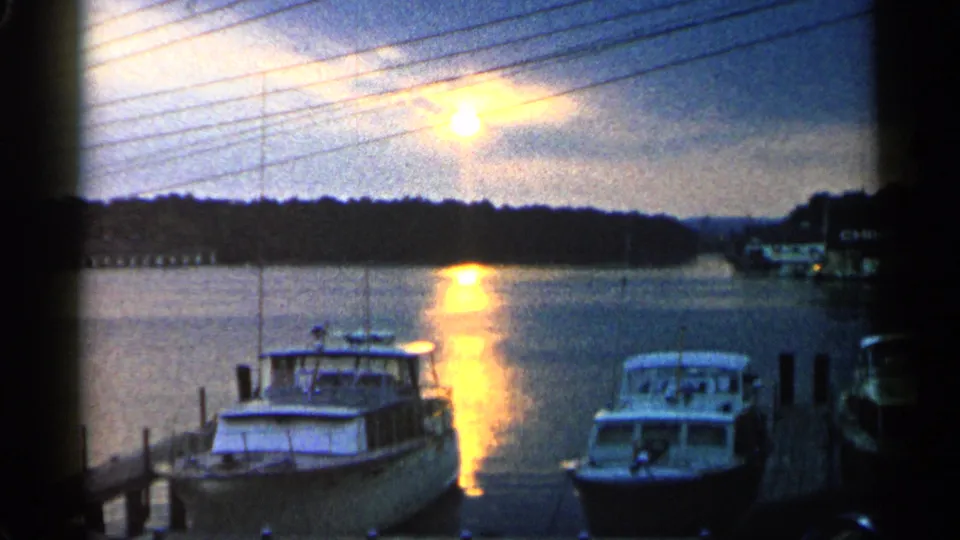 1960 Michigan marina frame with white cabin boats, dark shoreline, overhead utility lines, and sunset glare on the water.
