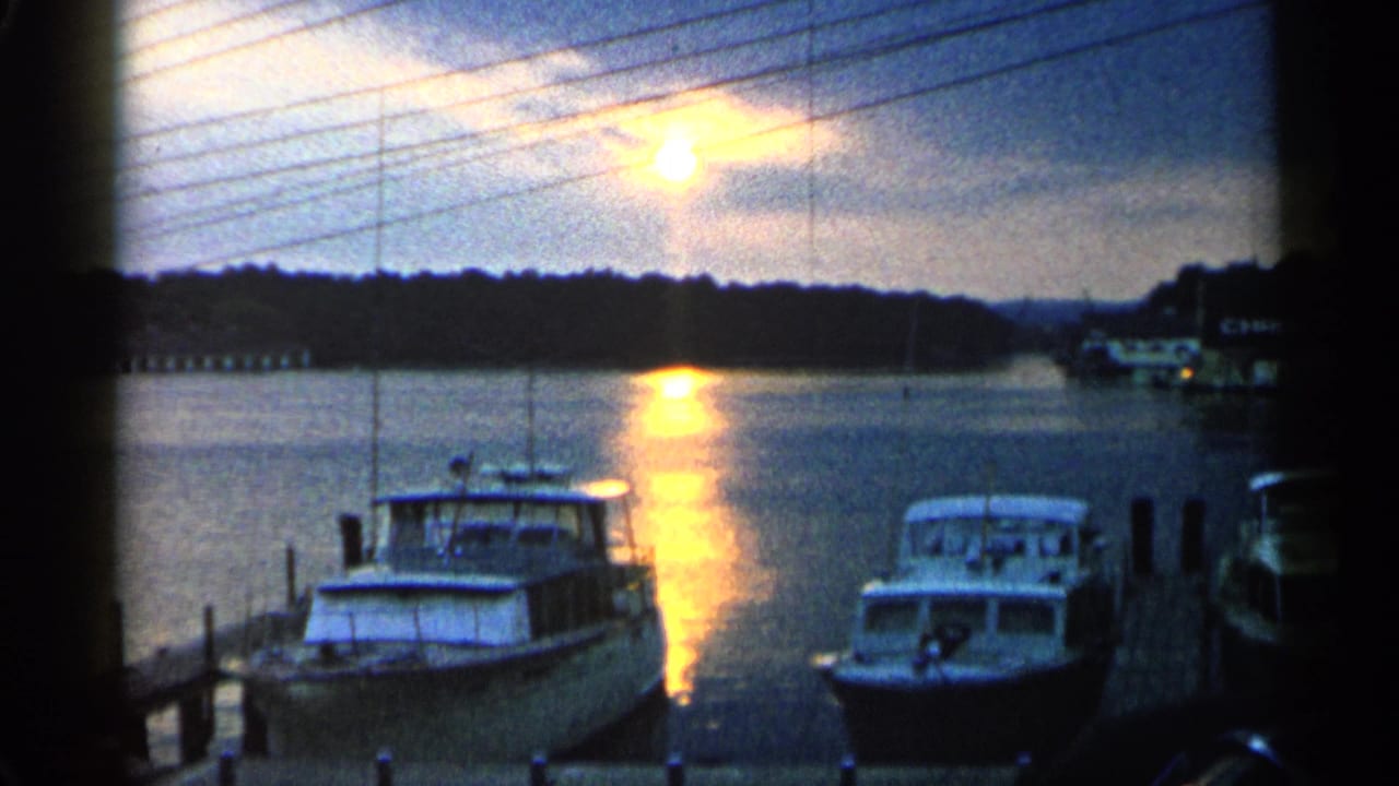 1960 Michigan home-movie frame showing docked boats on reflective water at sunset, with shoreline in the distance.
