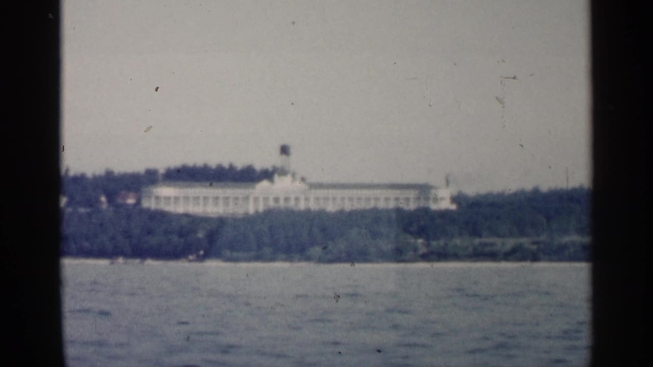 1960 Michigan home-movie frame looking across water toward a large pale building on a wooded shoreline.