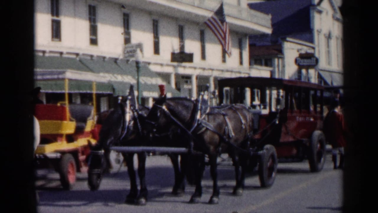 1960 Michigan home-movie frame showing horse-drawn carriages passing storefronts on a busy street with an American flag overhead.