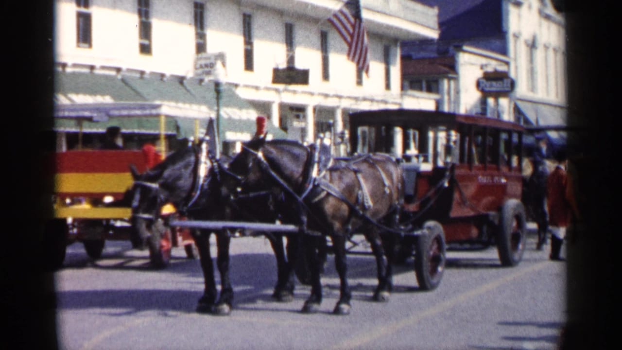 Horse team and red carriage pass along a Michigan town street, with storefront awnings, flags, and pedestrians visible in the background.