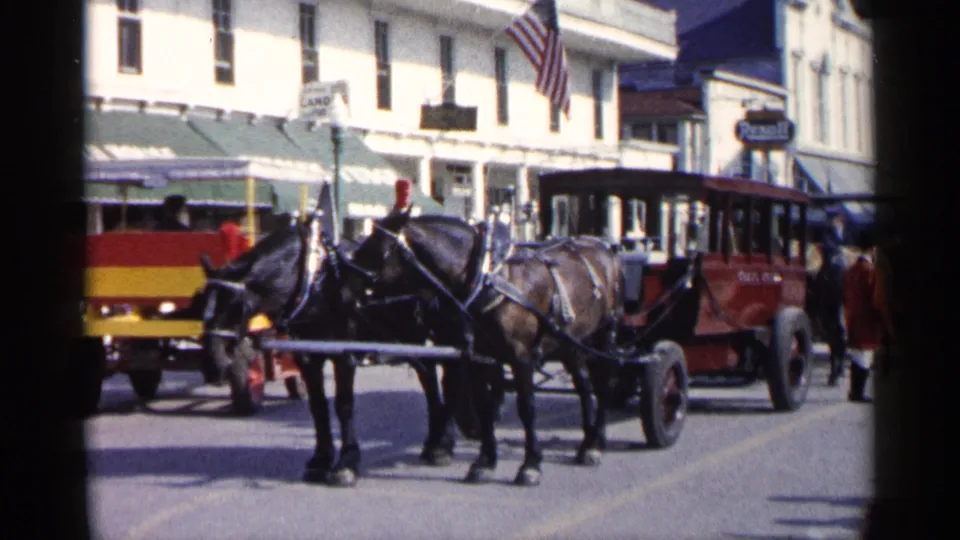 Horse team and red carriage pass along a Michigan town street, with storefront awnings, flags, and pedestrians visible in the background.