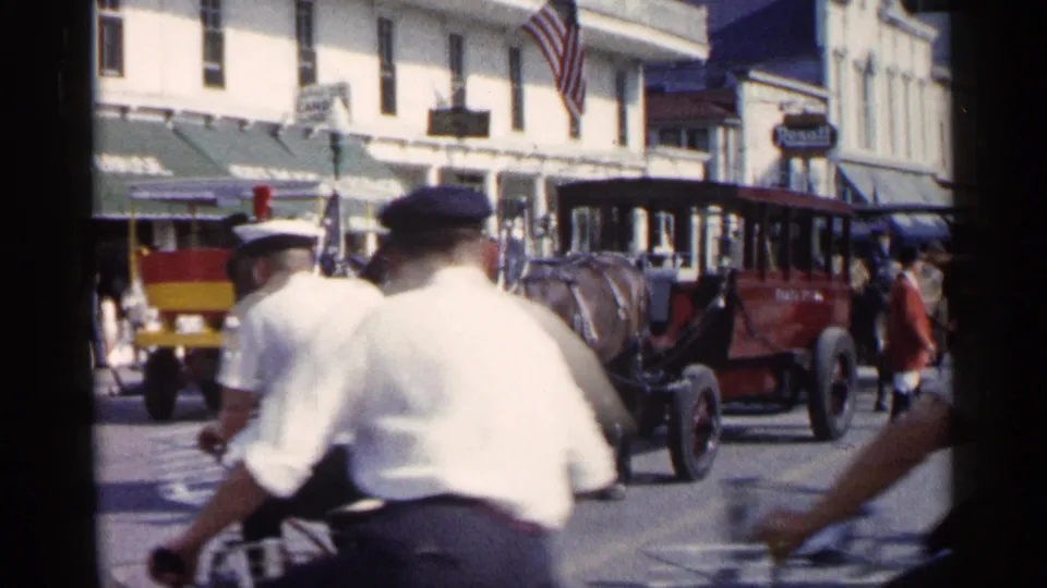 Cyclists in white shirts and caps ride across the foreground as horse-drawn carriages continue along the 1960 Michigan street scene.