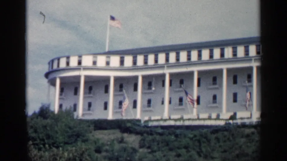 Wide 1960 Michigan home-movie view of a large white columned hotel exterior, likely the Grand Hotel, with multiple American flags flying.