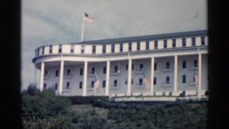 1960 Michigan home-movie frame showing a grand white civic-style building with columns, roof flag, front flags, cloudy sky, and foliage.