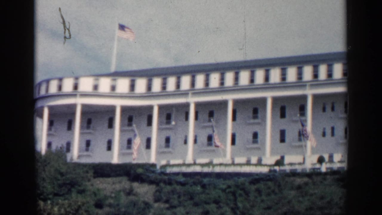 1960 Michigan frame of the same large white civic-style facade, with American flags along the front and a flagpole above the roofline.