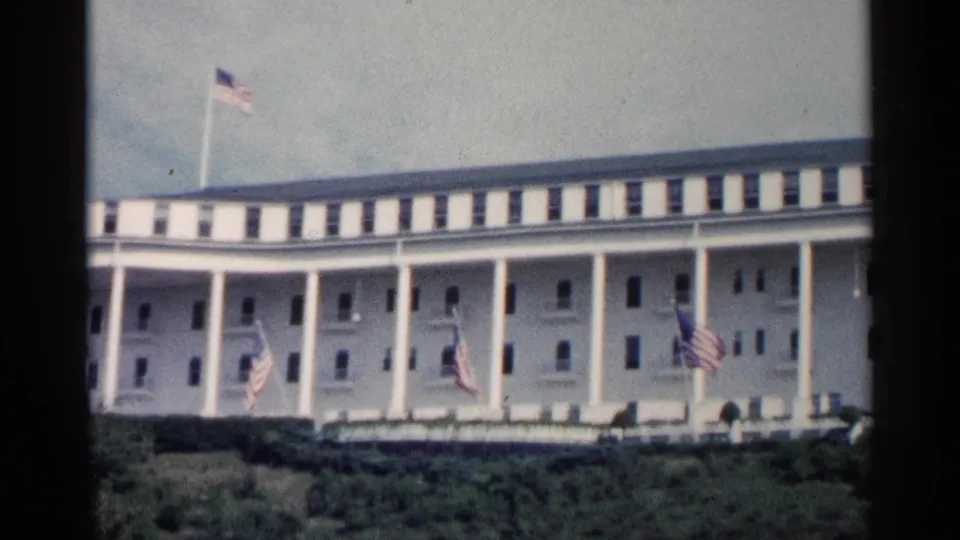 1960 Michigan home-movie frame with the building slightly closer in view, showing columns, multiple American flags, windows, and green landscaping.