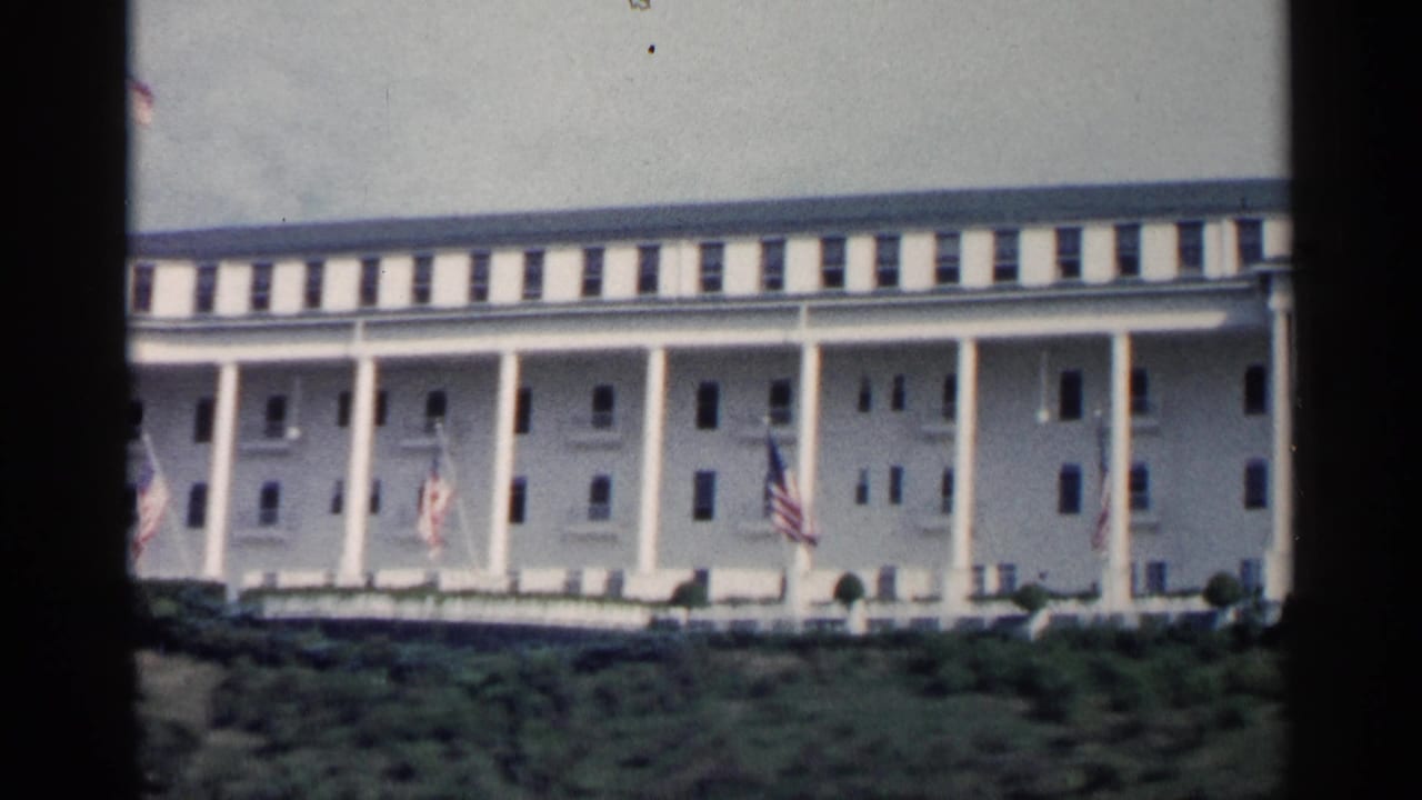 1960 Michigan 8mm view of a grand white colonnaded building with American flags and low greenery in front.