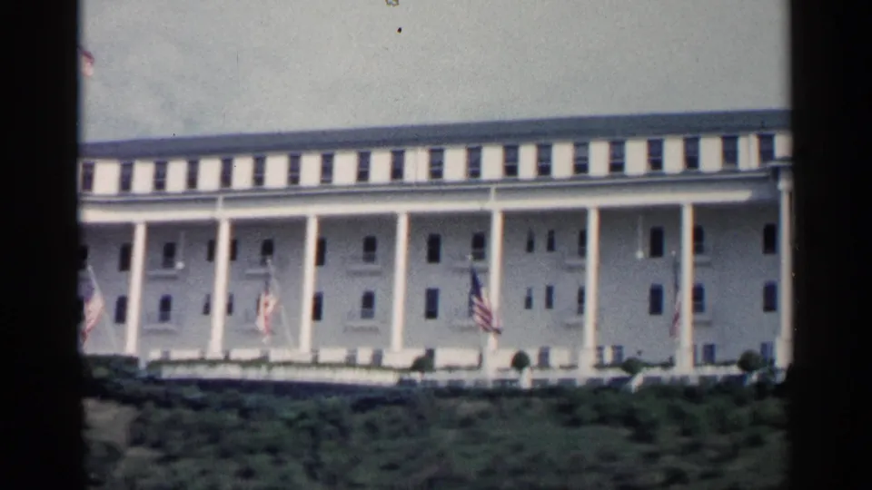 1960 Michigan 8mm view of a grand white colonnaded building with American flags and low greenery in front.