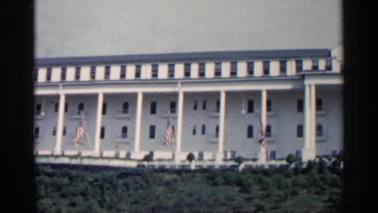 White colonnaded building facade with flags and shrubs in the foreground.