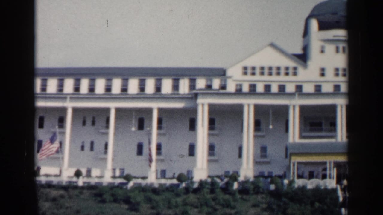 1960 Michigan home-movie frame showing a white resort-style building with tall columns, a green dome, American flag, shrubs, and visitors outside.