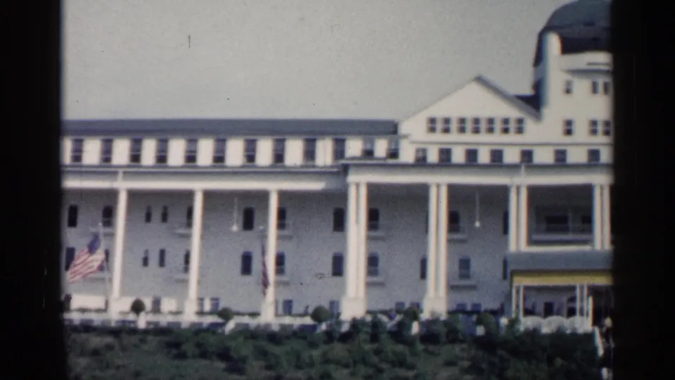 1960 Michigan home-movie frame showing a white resort-style building with tall columns, a green dome, American flag, shrubs, and visitors outside.
