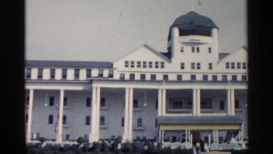 Closer 1960 Michigan home-movie frame of the white resort facade, showing the green dome, upper windows, tall columns, shaded porch, and visitors by the entrance.