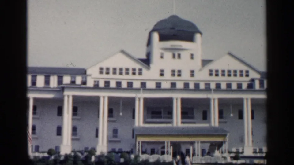 1960 Michigan frame showing the white resort-style facade with green dome, rows of windows, columned porch, entrance canopy, flags, shrubs, and visitors outside.