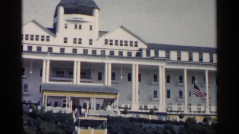 1960 Michigan home-movie frame of the resort front with tall white columns, entrance canopy, rows of windows, visitors, and an American flag at right.