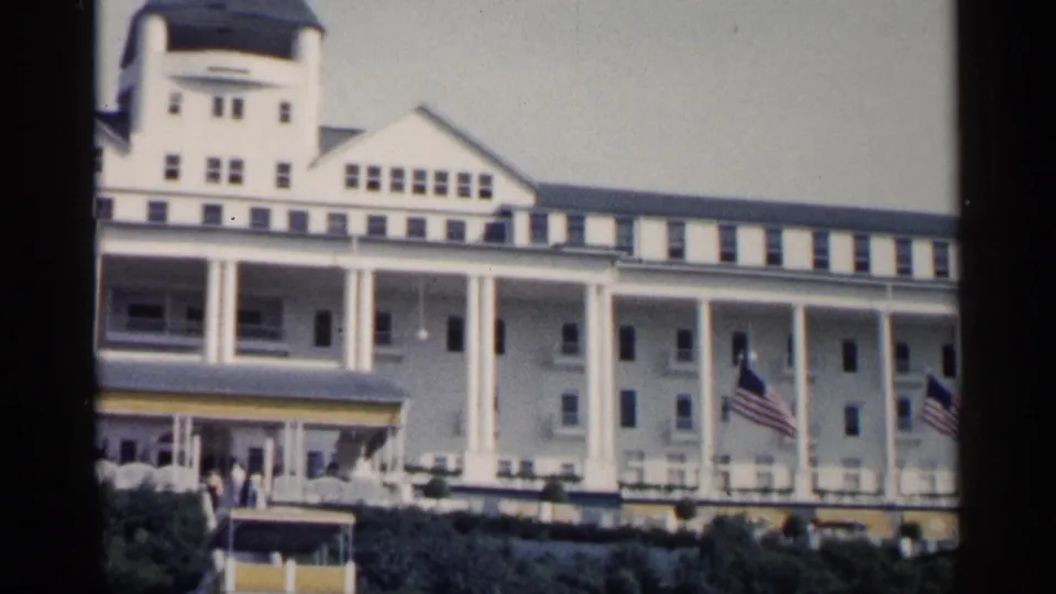 1960 Michigan 8mm frame showing the white resort's long columned porch, partial entrance canopy, distant visitors, landscaping, and American flags at right.