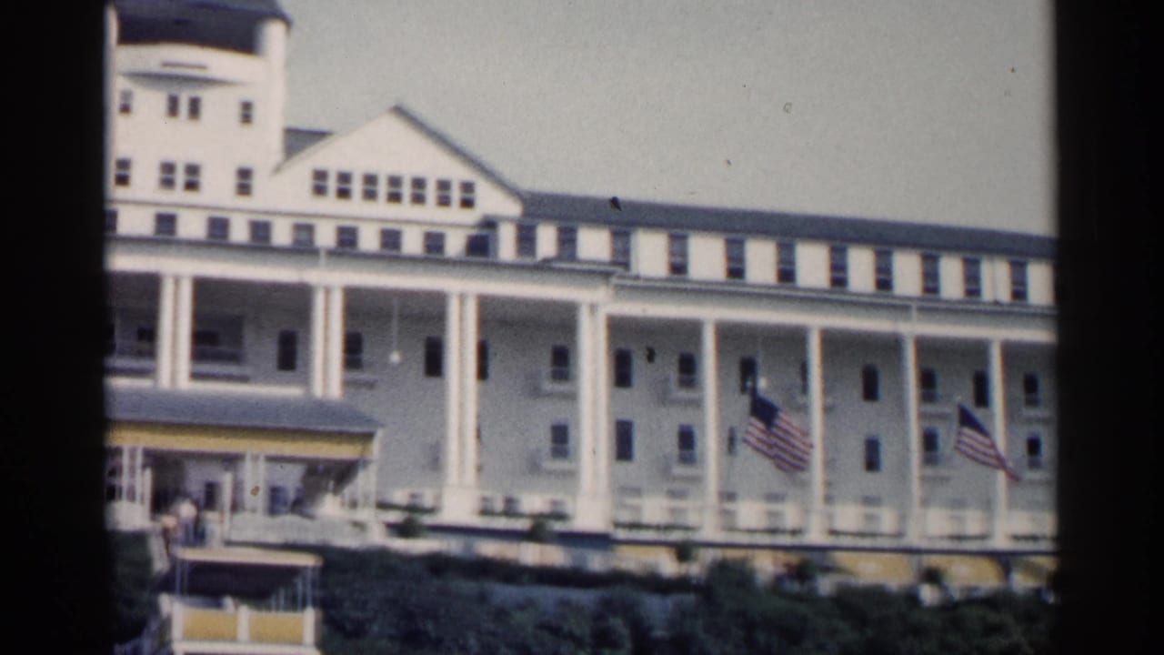 1960 Michigan 8mm frame showing a large white resort hotel with tall columns, dormer windows, flags, awnings, and shrubs.