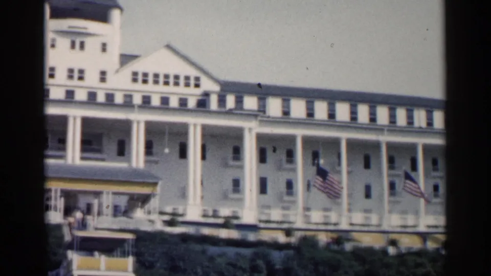 1960 Michigan 8mm frame showing a large white resort hotel with tall columns, dormer windows, flags, awnings, and shrubs.