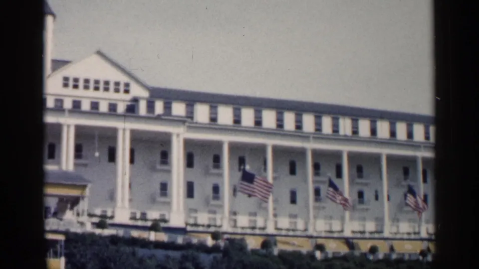 The camera view continues across the 1960 Michigan resort exterior, showing the colonnade, rows of windows, and multiple American flags.