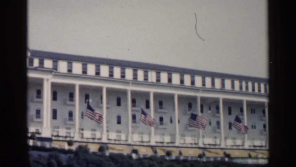 The final frame holds on the white Michigan resort exterior, with repeated porch columns, flags, awnings, and shrubs under a bright sky.