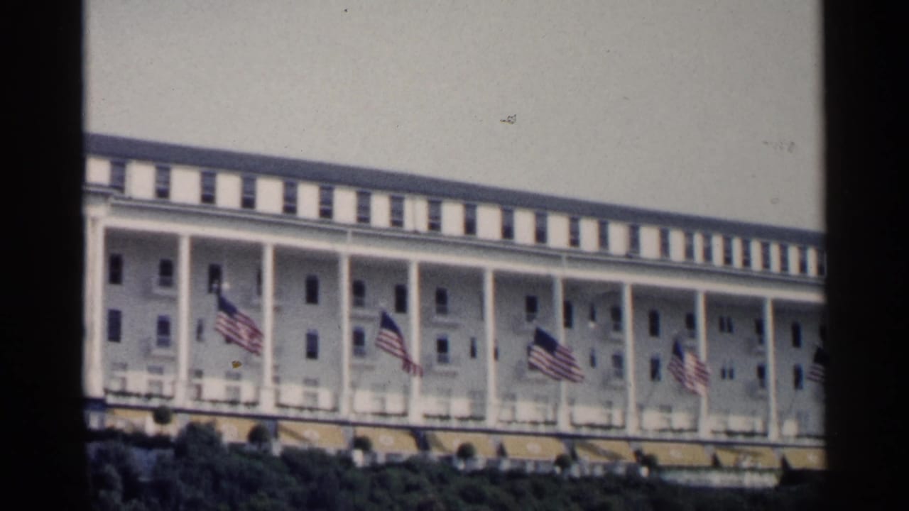 1960 Michigan home-movie view of a long white resort hotel exterior with columns, striped awnings, and several American flags.
