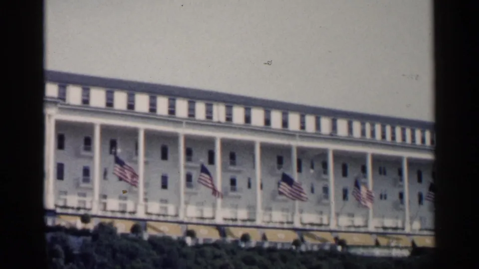 1960 Michigan home-movie view of a long white resort hotel exterior with columns, striped awnings, and several American flags.