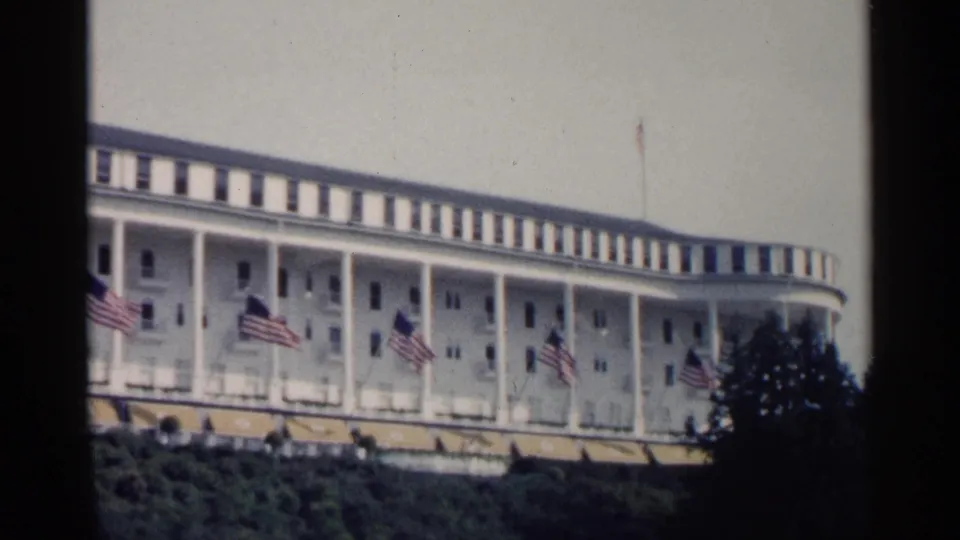 1960 Michigan resort footage frames the white hotel exterior, flags, awnings, and landscaped foreground in soft 8mm color.