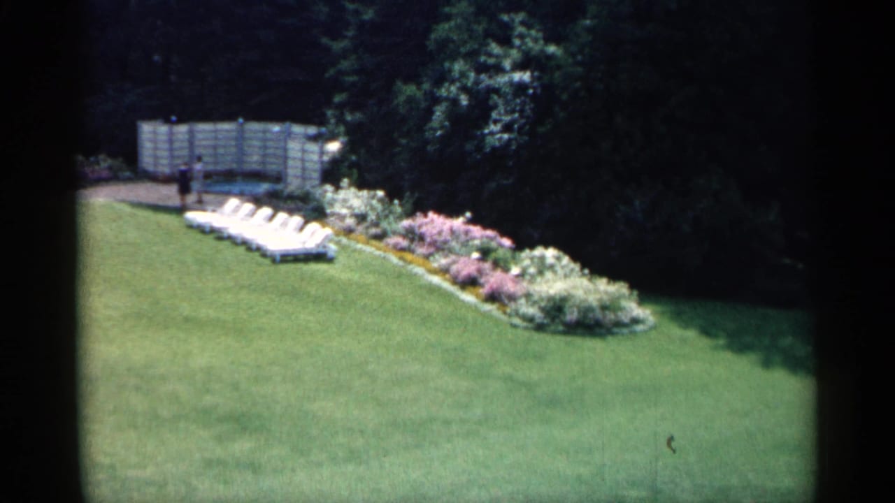 1960 Michigan home-movie frame showing a suburban lawn, flowering border, white lounge chairs, fence, trees, and a distant person.