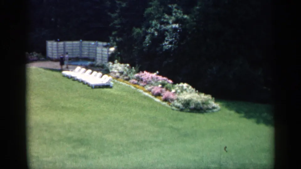1960 Michigan home-movie frame showing a suburban lawn, flowering border, white lounge chairs, fence, trees, and a distant person.