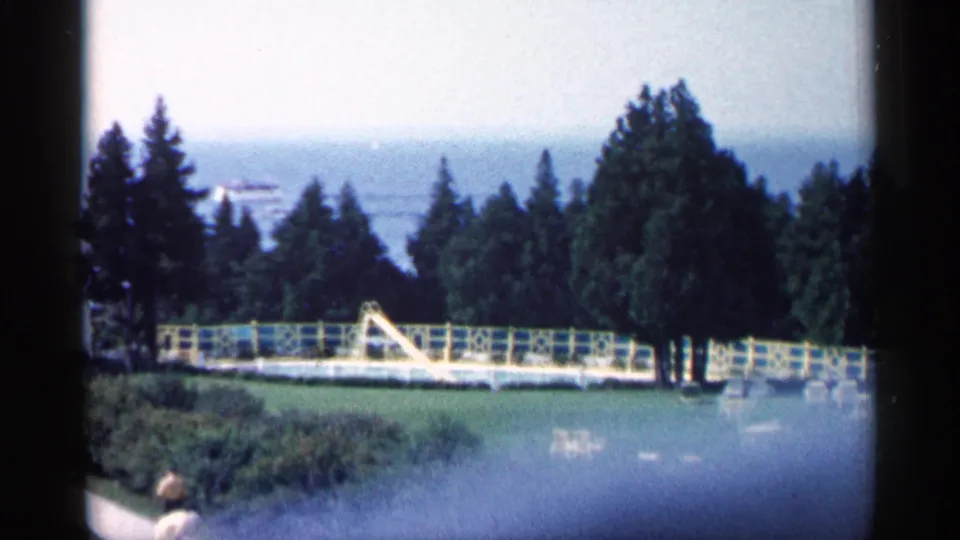 1960 Michigan home-movie frame showing a pool area with yellow slide, fence, trees, distant water, and a partial foreground figure.