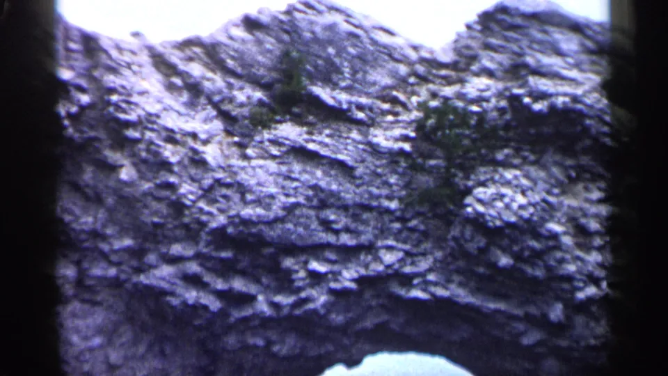 1960 Michigan 8mm frame showing textured rock layers, sparse greenery, and a pale foreground edge near the bottom.