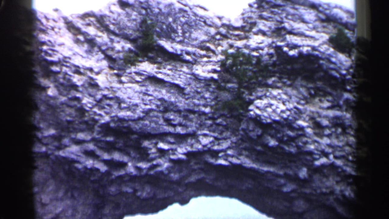 1960 Michigan 8mm frame showing rough eroded rock layers, small vegetation, and a bright natural arch opening at the lower edge.