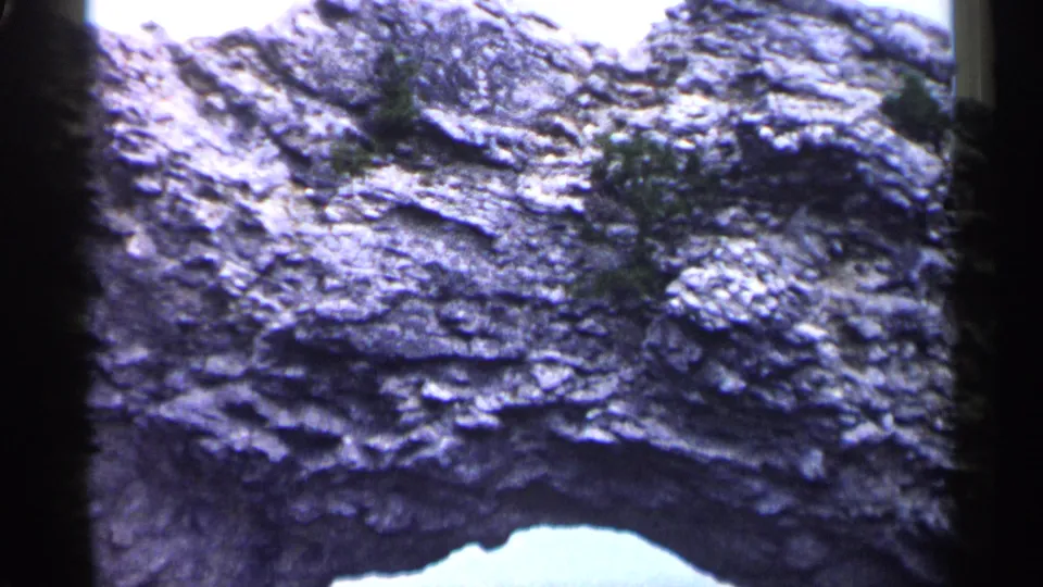 1960 Michigan 8mm frame showing rough eroded rock layers, small vegetation, and a bright natural arch opening at the lower edge.