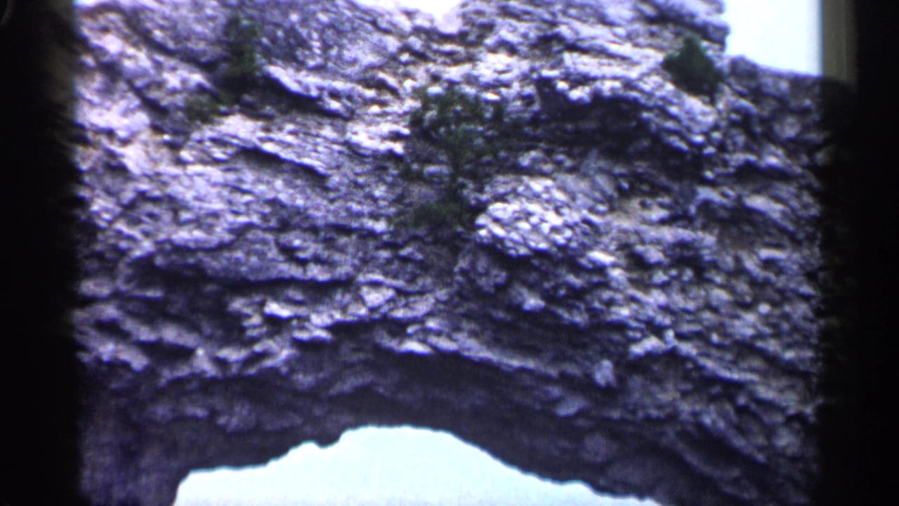 1960 Michigan 8mm frame holding on a rugged rock face with a light-filled natural arch opening and sparse vegetation.