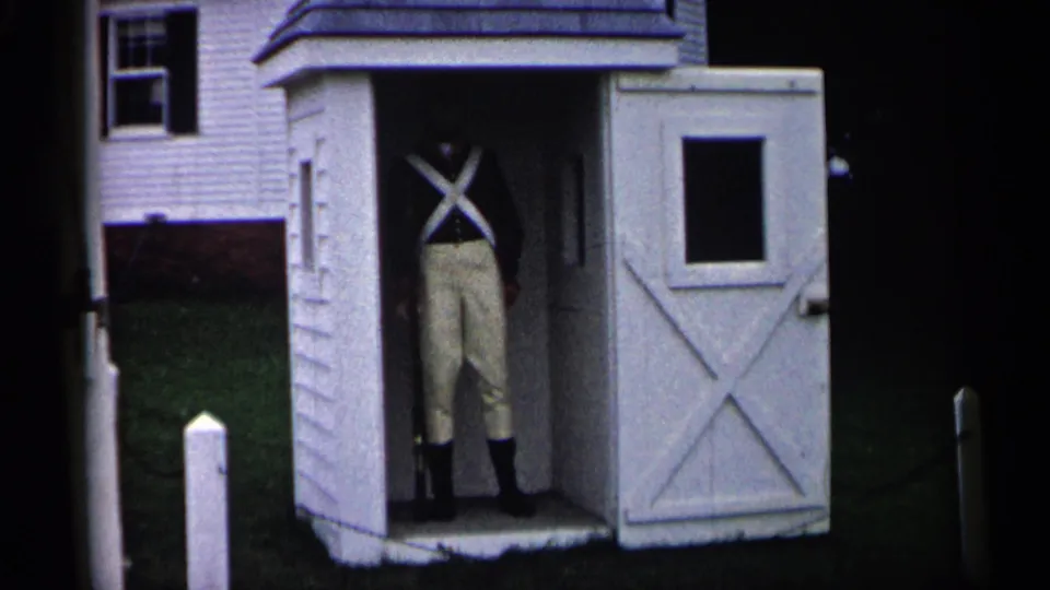 1960 Michigan home-movie frame of a visible adult in colonial-style dress inside a small white reenactment structure.
