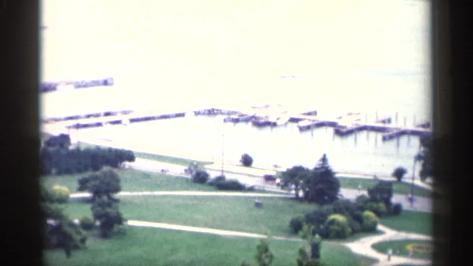 1960 Michigan 8mm frame looking over green lawns, paths, trees, docks, and a bright waterfront area.