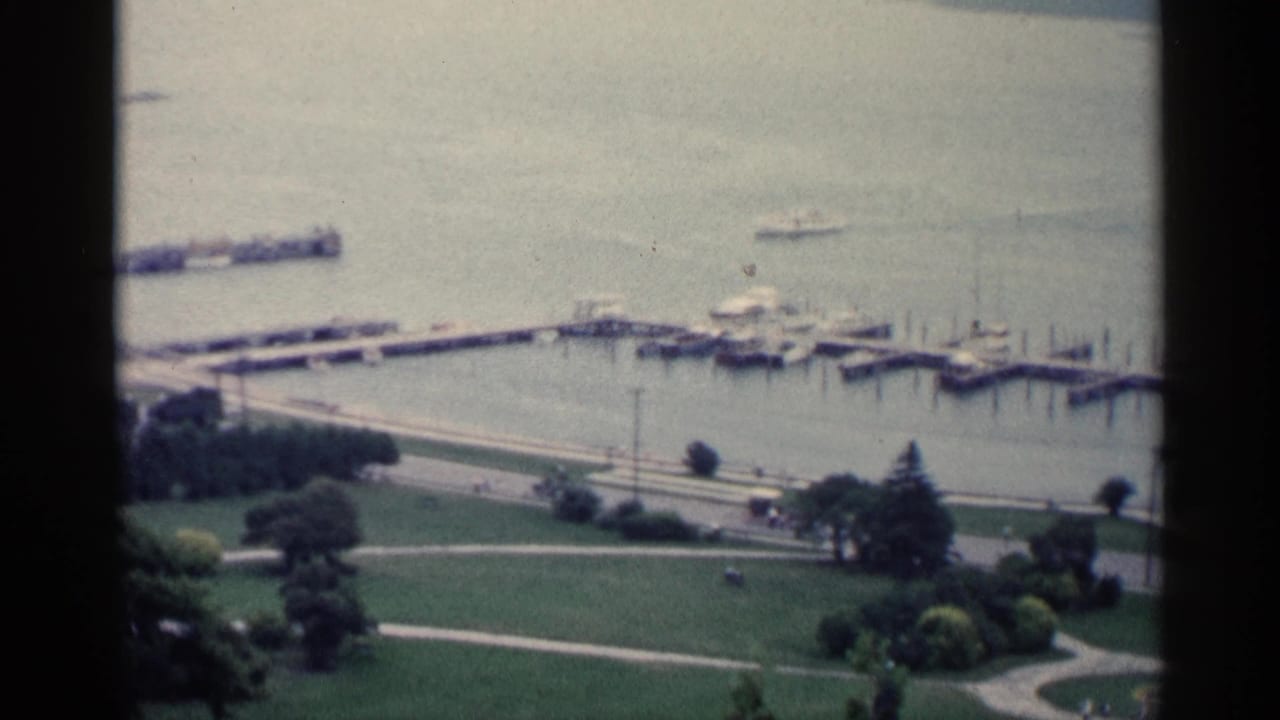 Elevated view of a bay with docks, small boats, lawns, paths, and trees along the waterfront.