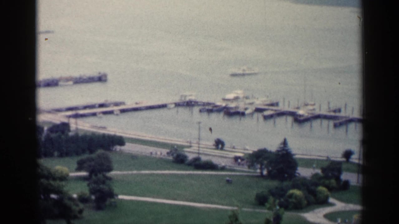 1960 Michigan lakeside view with docks, small boats, green parkland, and distant people along the waterfront promenade.
