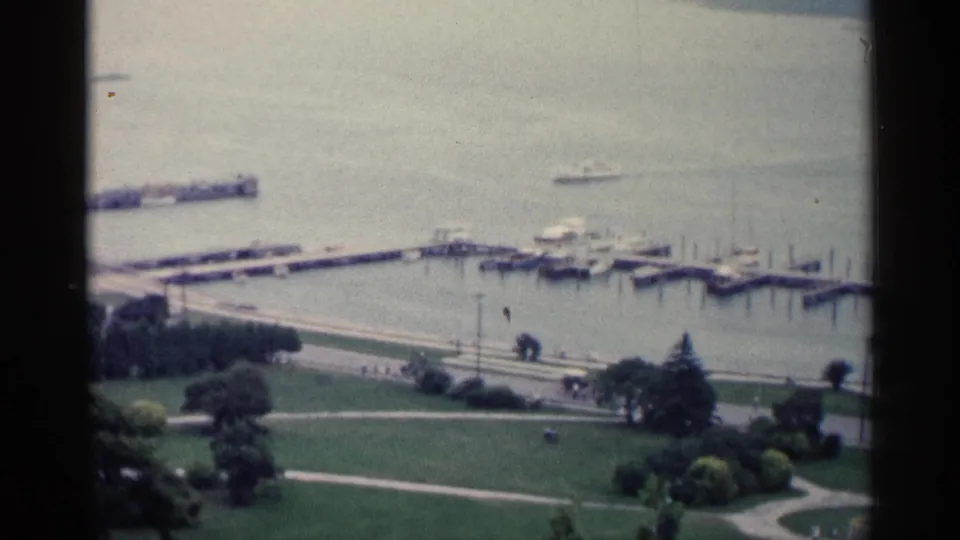 1960 Michigan lakeside view with docks, small boats, green parkland, and distant people along the waterfront promenade.