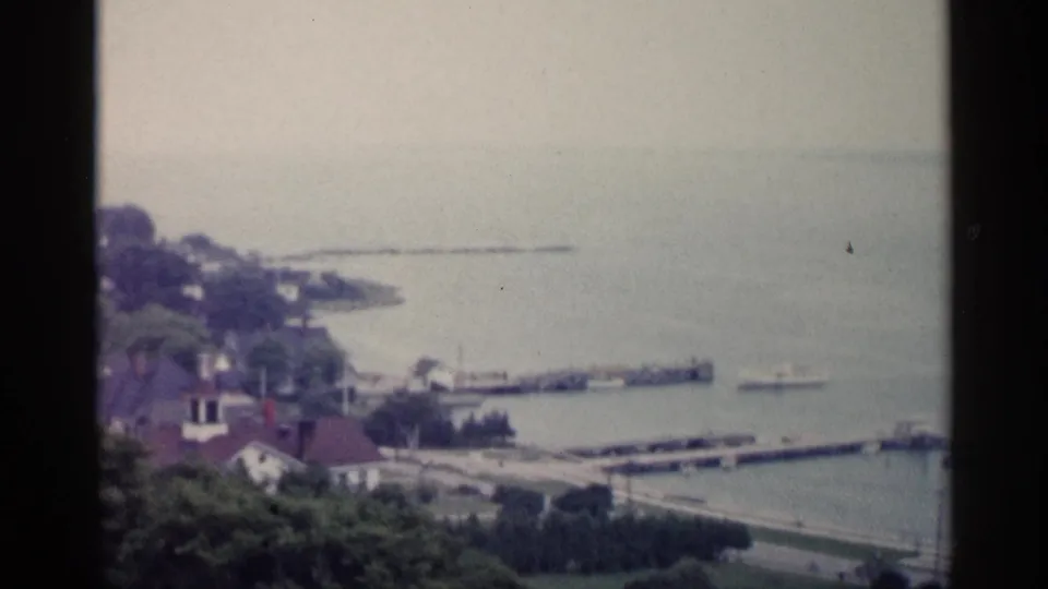 1960 Michigan home-movie frame with shoreline residences, marina docks, small boats, trees, and pale bay water in soft focus.